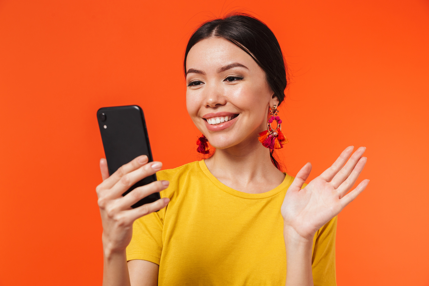 Happy Young Woman Posing Isolated over Orange Wall Background Talking by Mobile Phone Take a Selfie Waving.